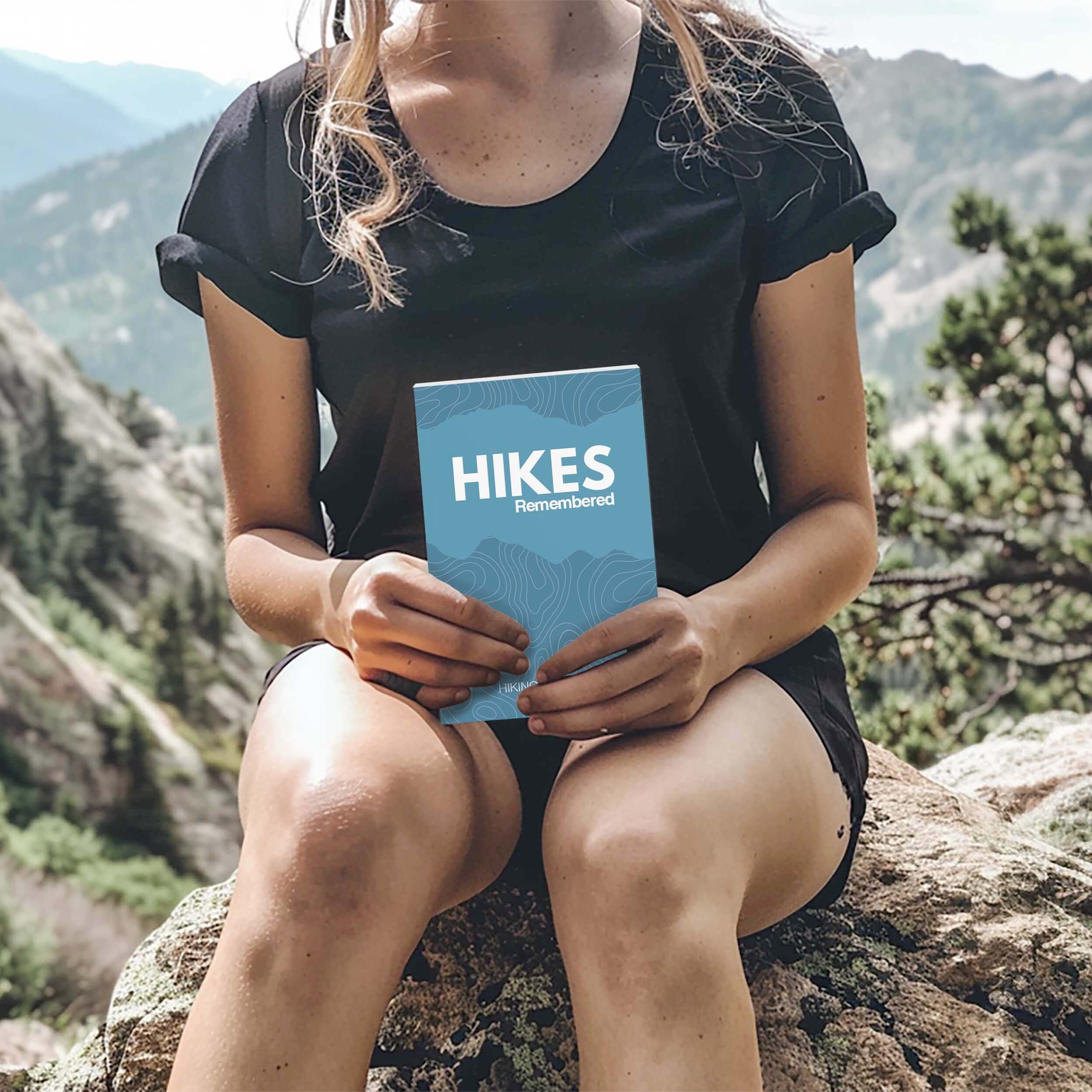 Person holding the Hikes Remembered logbook journal in a scenic mountain landscape.